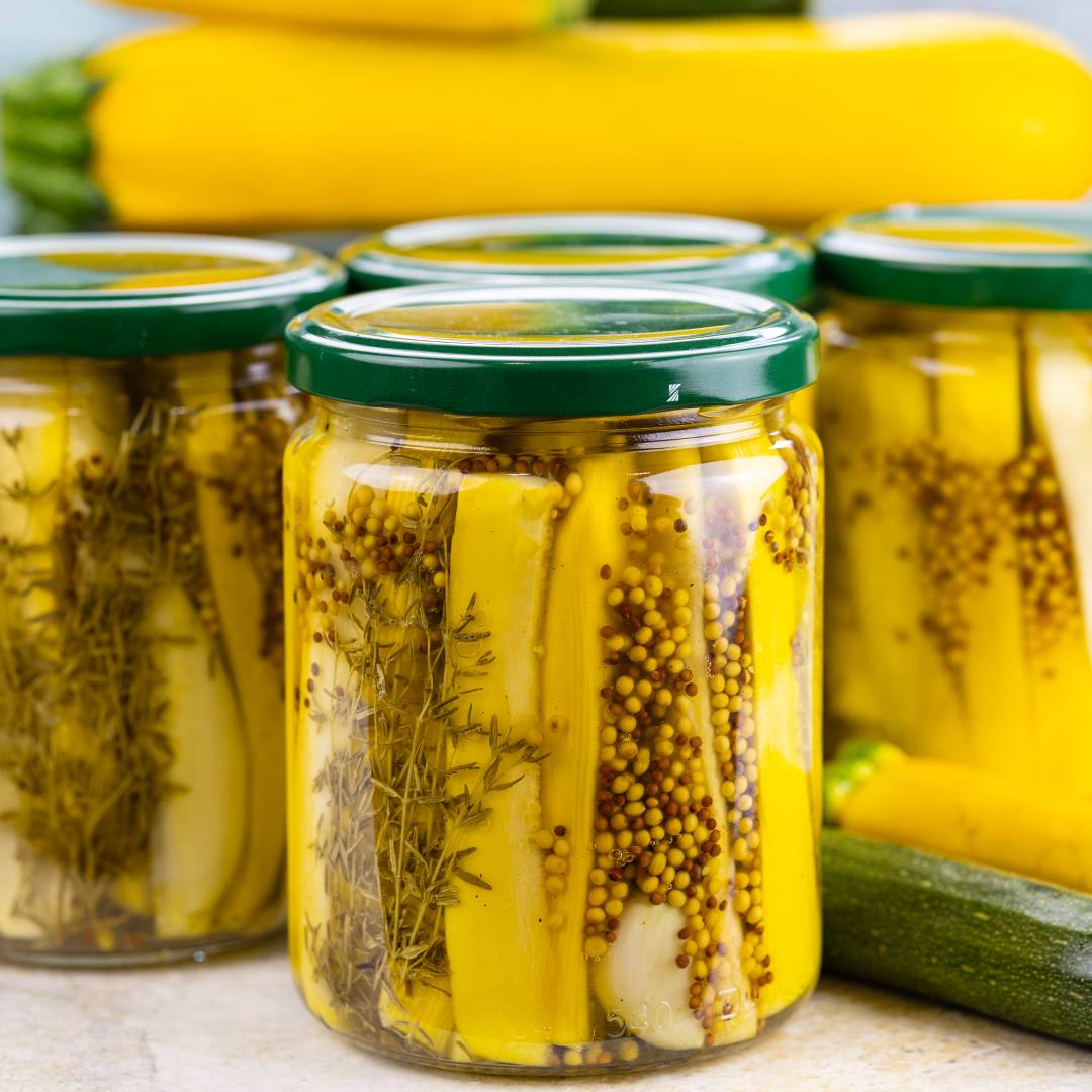 Szklane słoiki z zielonymi zakrętkami wypełniają żółte słupki cukinii. W przezroczystej zalewie zanurzone są ziarenka musztardy francuskiej, gałązki świeżego tymianku oraz ząbki czosnku. W tle leżą całe żółte i zielone warzywa.