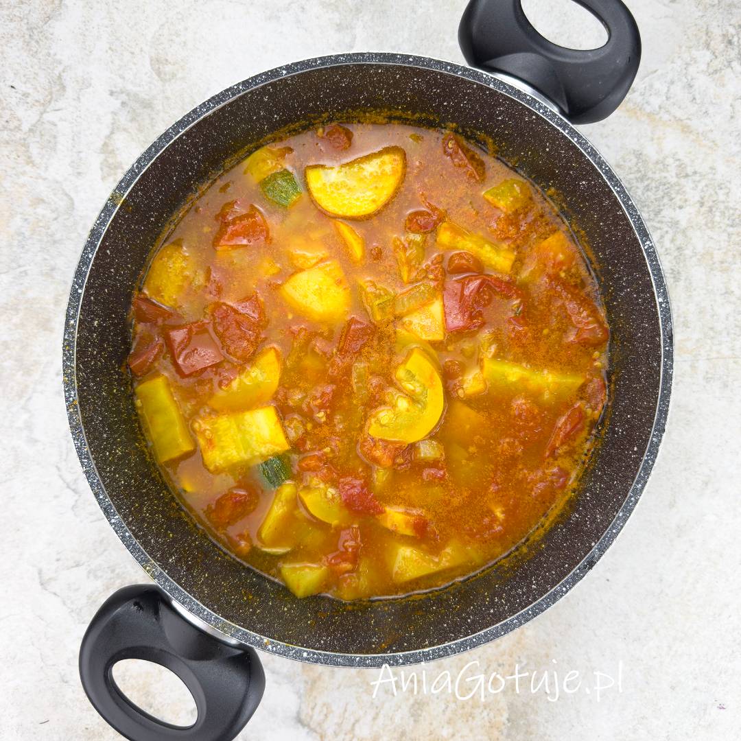 Zupa z cukinii i pomidorów podczas gotowania w ciemnym garnku o nakrapianej fakturze. W żółtawo pomarańczowym bulionie z dodatkiem curry znajdują się miękkie kawałki żółtej i zielonej cukinii, cząstki czerwonych pomidorów oraz ziemniaków.