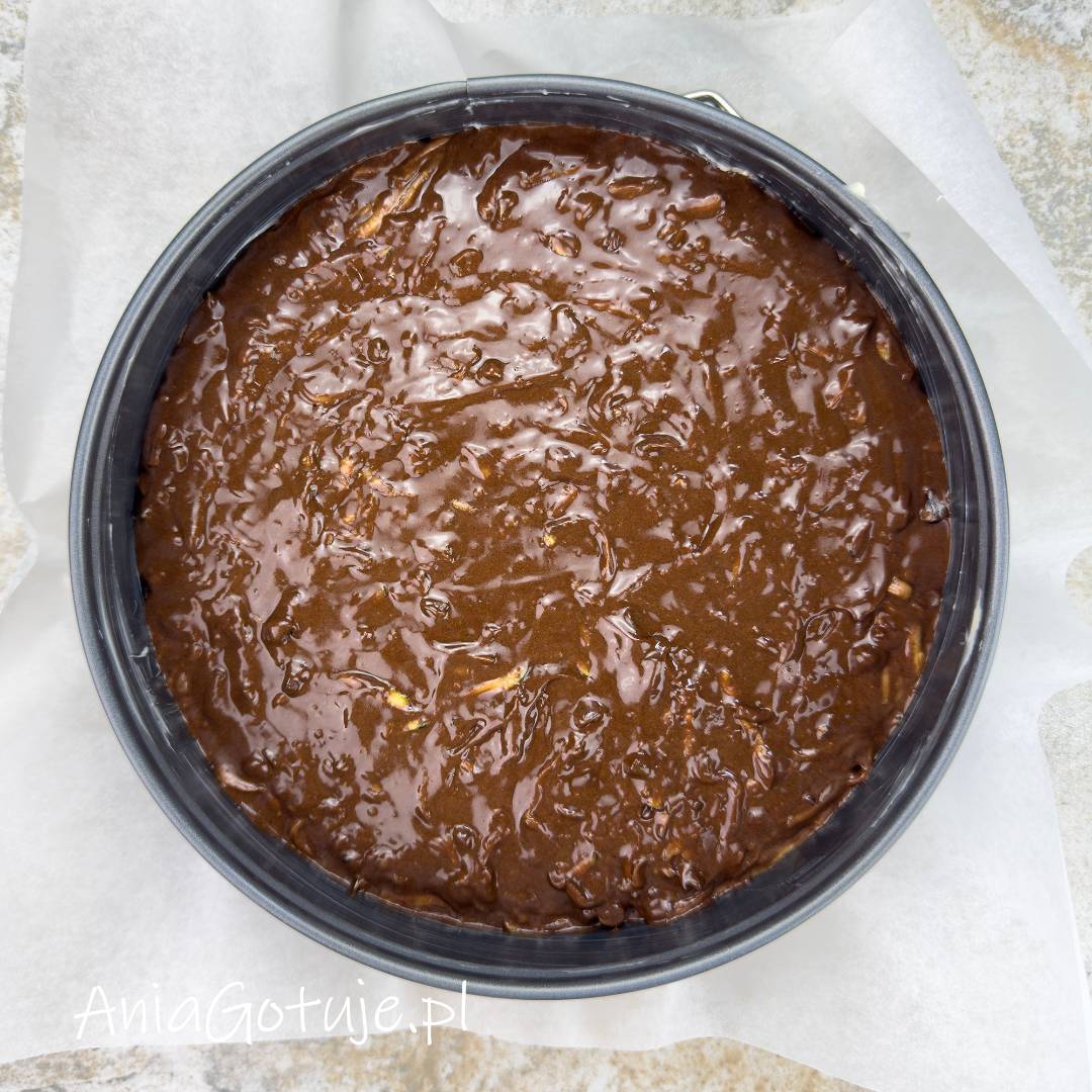 Gęste, klejące ciasto brownie wypełnia okrągłą formę wyłożoną papierem do pieczenia. Ciemna, czekoladowa masa ma lśniącą, lekko nieregularną powierzchnię, która została rozprowadzona po całym dnie.