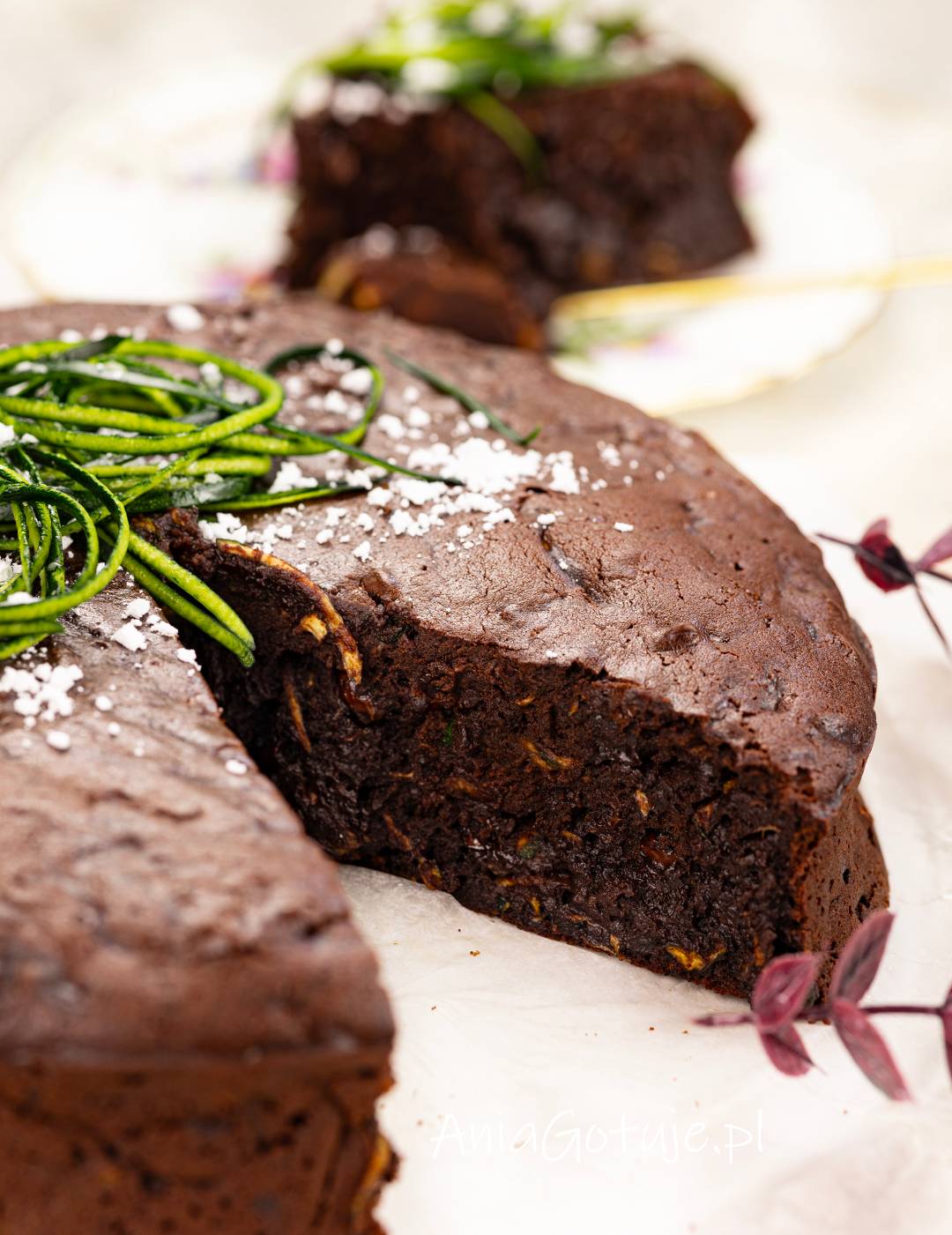 Upieczone brownie ma ciemną, lekko popękaną powierzchnię i gęsty, wilgotny środek z widocznymi kawałkami cukinii. Wierzch posypano białym cukrem pudrem i udekorowano długimi zielonymi paskami cukinii.