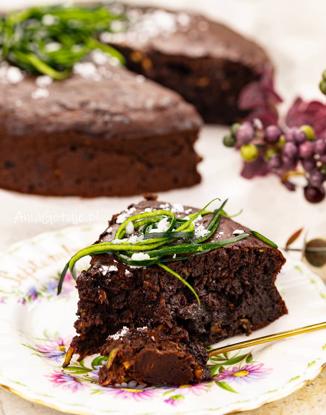 Gruby kawałek brownie ma niemal czarny, wilgotny środek z kawałkami czekolady i znajduje się na dekoracyjnym talerzu. Wierzch zdobi cukier puder oraz długie, zielone paski cukinii, obok umieszczono złoty widelec, a w tle znajduje się reszta ciasta.