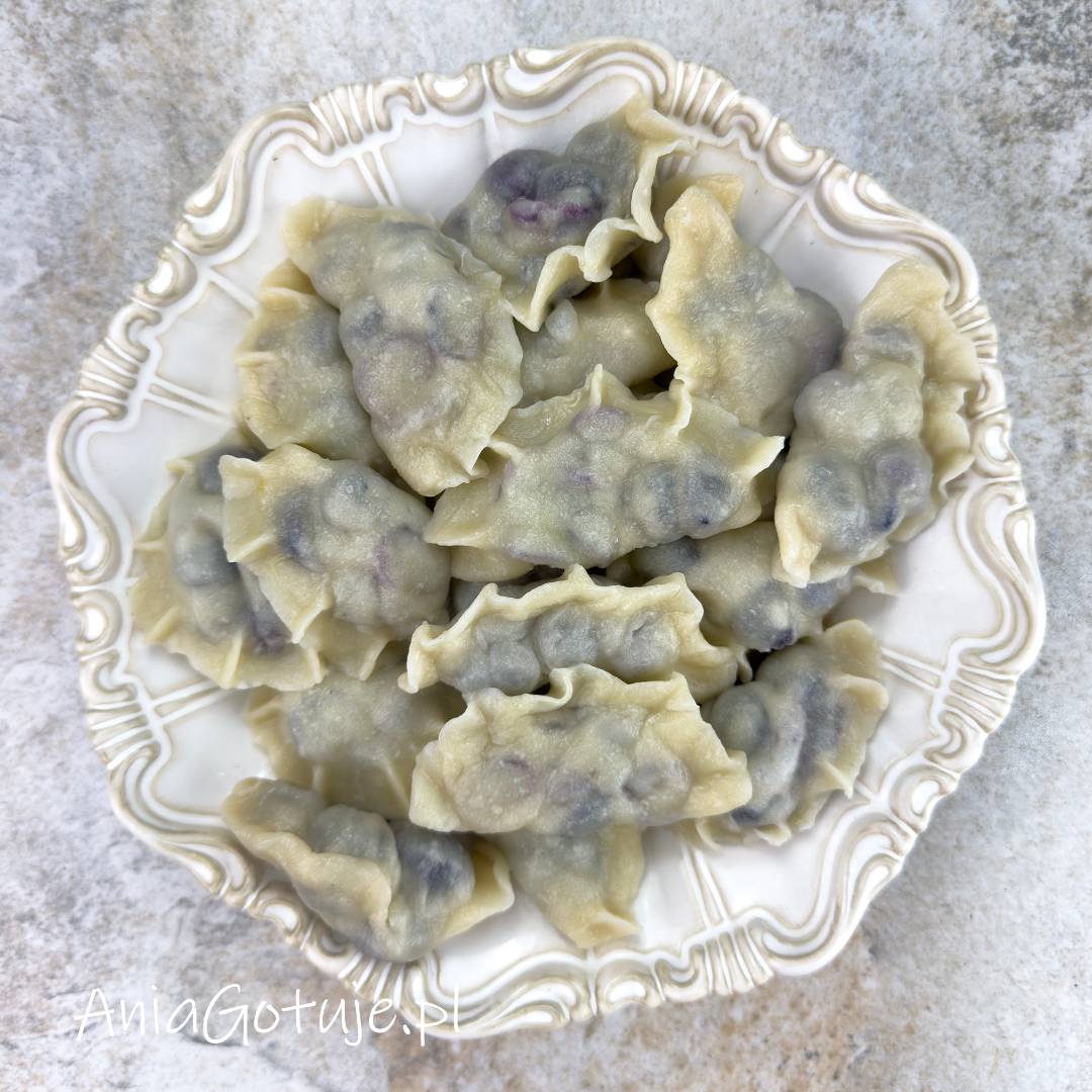 Ugotowane pierogi z borówkami są ułożone na białym talerzu z ozdobnymi złotymi detalami. Jasne, lekko półprzezroczyste ciasto ma pofałdowane brzegi, a fioletowe owoce prześwitują przez jego powierzchnię.
