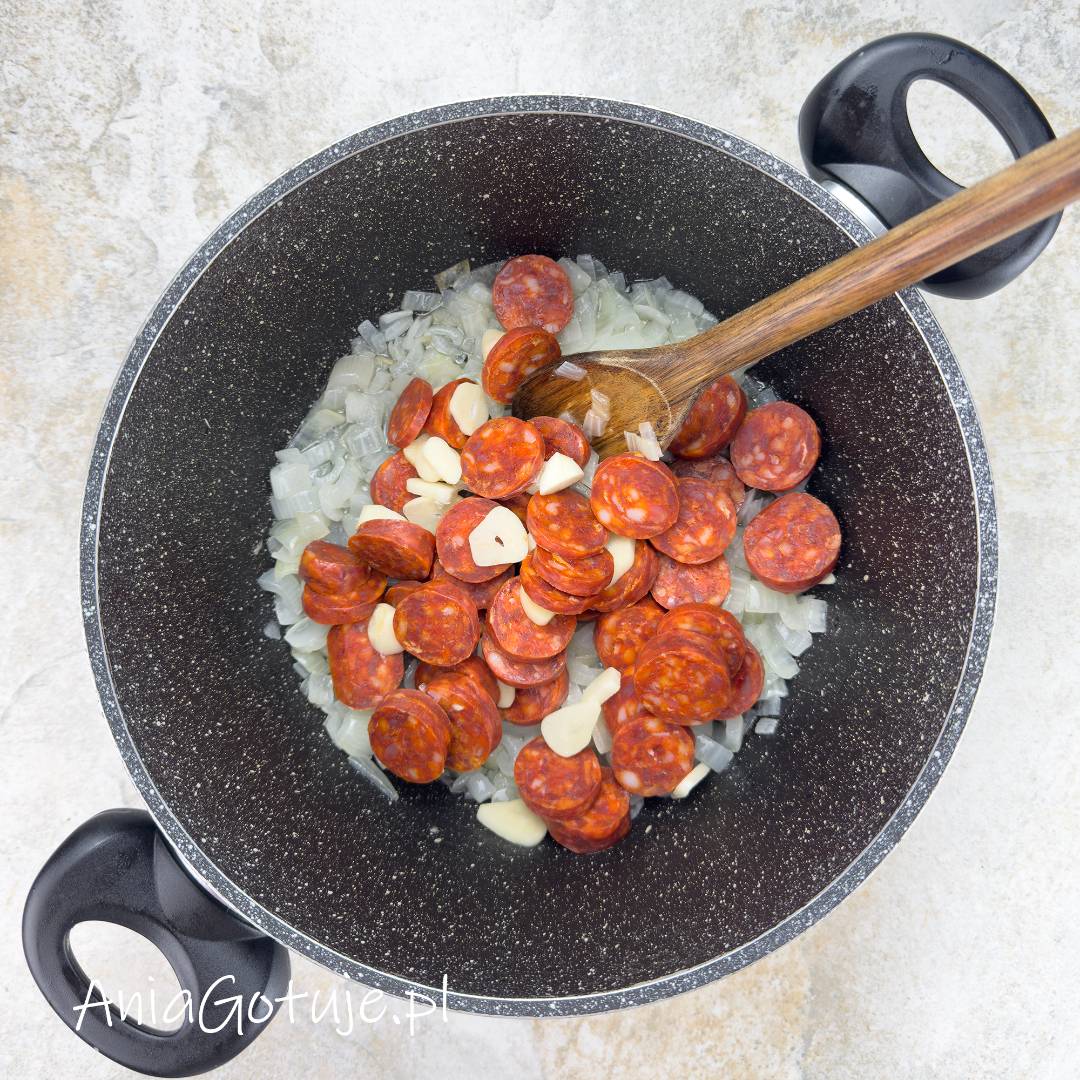 W szerokim czarnym garnku znajduje się warstwa posiekanej cebuli, na której ułożono czerwone plastry chorizo oraz jasne plasterki czosnku. Drewniana łyżka jest umieszczona nad składnikami i służy do ich wymieszania podczas smażenia.
