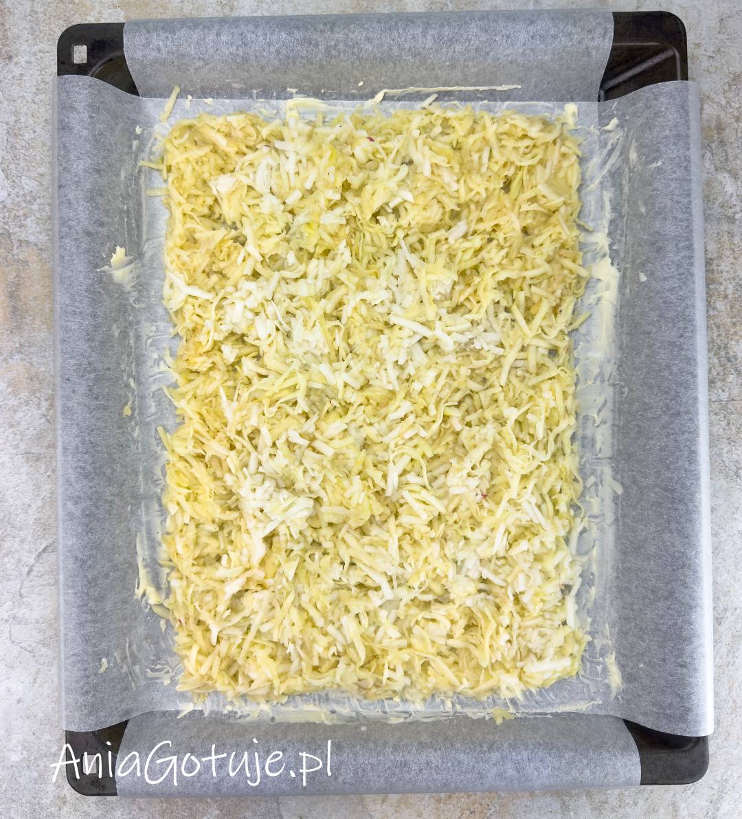 Głęboka blacha jest wyłożona papierem do pieczenia, którego brzegi wystają poza formę. W środku znajduje się jasna, lekko wilgotna warstwa startych jabłek, rozprowadzona w równy prostokąt. Masło pokrywa papier przy brzegach.