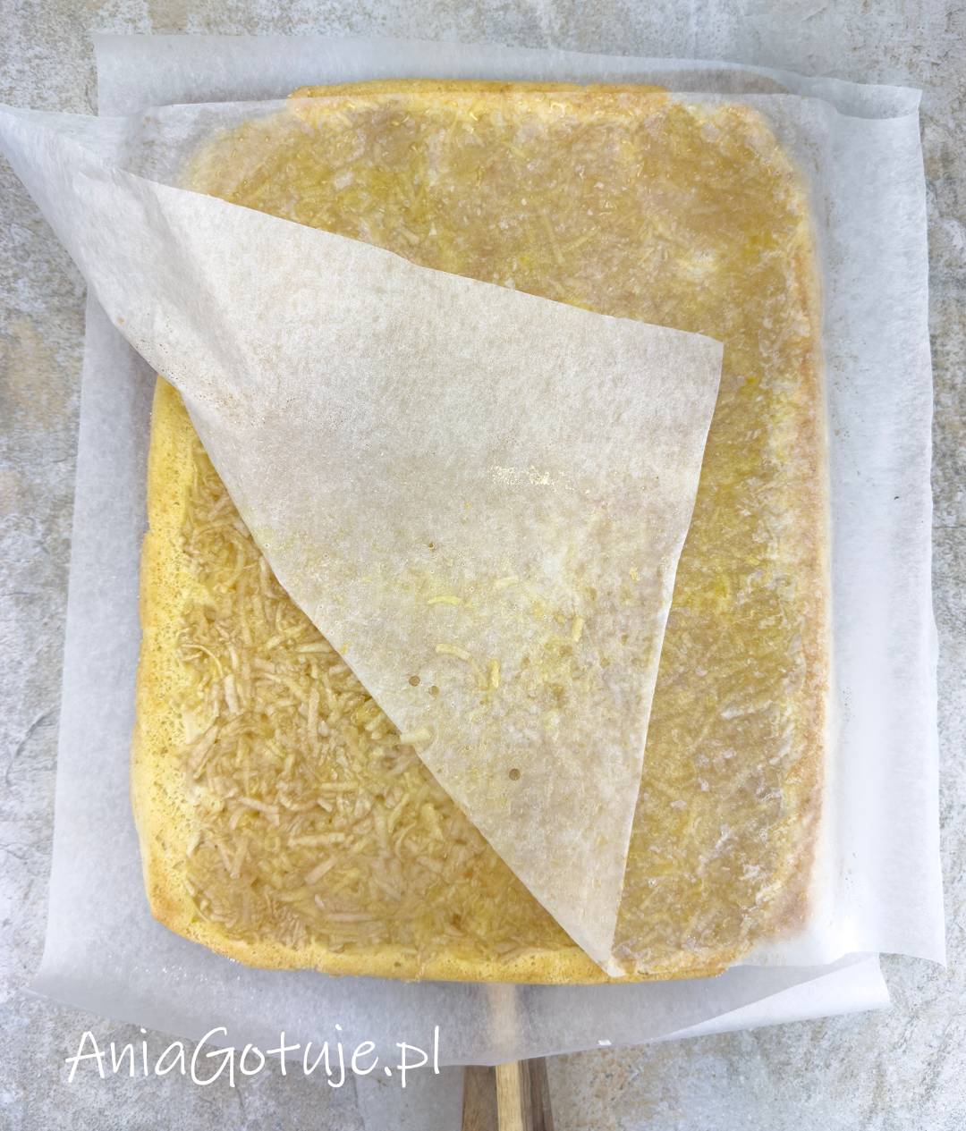 Ciepły, prostokątny biszkopt ma na wierzchu jasną warstwę startych jabłek. Papier do pieczenia jest delikatnie odciągany po skosie, a ciasto pozostaje ułożone na świeżym arkuszu.