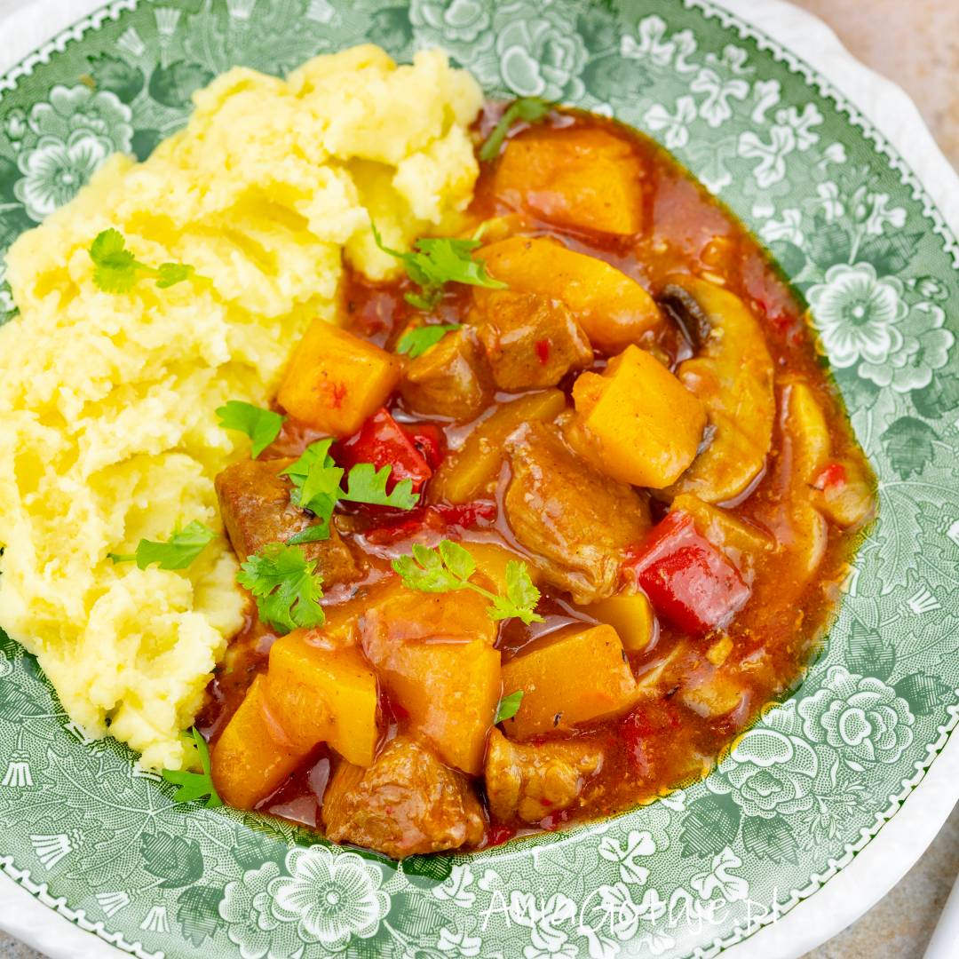 Gęsty gulasz w czerwono-brązowym sosie zawiera złote kawałki dyni, mięso, czerwoną paprykę i pieczarki. Porcja została ułożona obok jasnego puree ziemniaczanego na zielonym, wzorzystym talerzu i udekorowana zielonymi ziołami.
