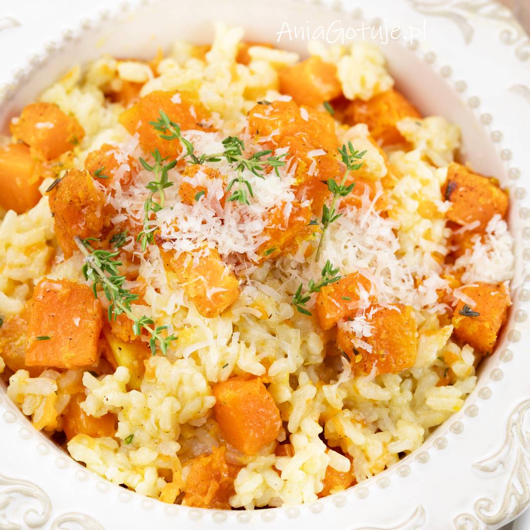 Jasne, kremowe ziarna ryżu są wymieszane z kawałkami pomarańczowej dyni i podane na białym, ozdobnym talerzu. Wierzch zdobi starty parmezan oraz świeże gałązki tymianku.