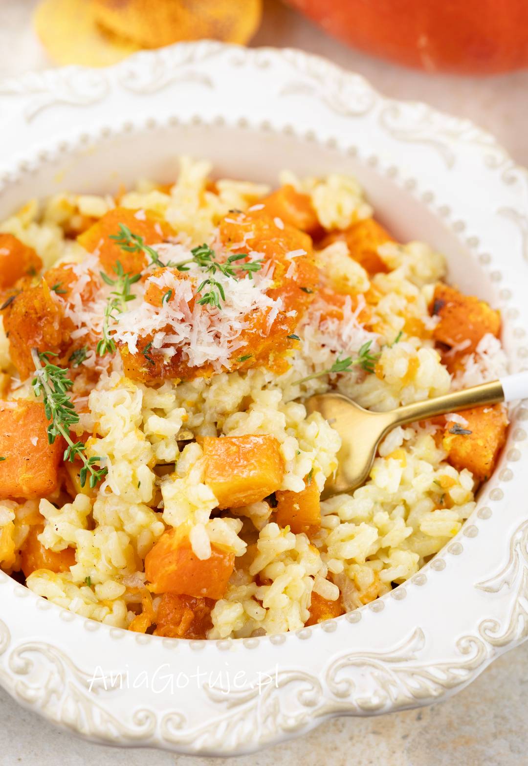 Jasne, kremowe risotto z dużymi kawałkami pomarańczowej dyni wypełnia białe, ozdobne naczynie. Na wierzchu znajdują się starty parmezan i zielone gałązki tymianku, a po prawej stronie umieszczona jest złota łyżka.